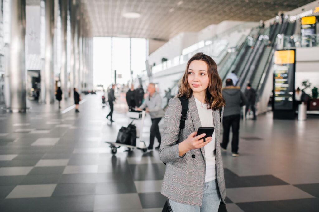 Frau mit Gepäck und Handy in der Hand steht am Flughafen.