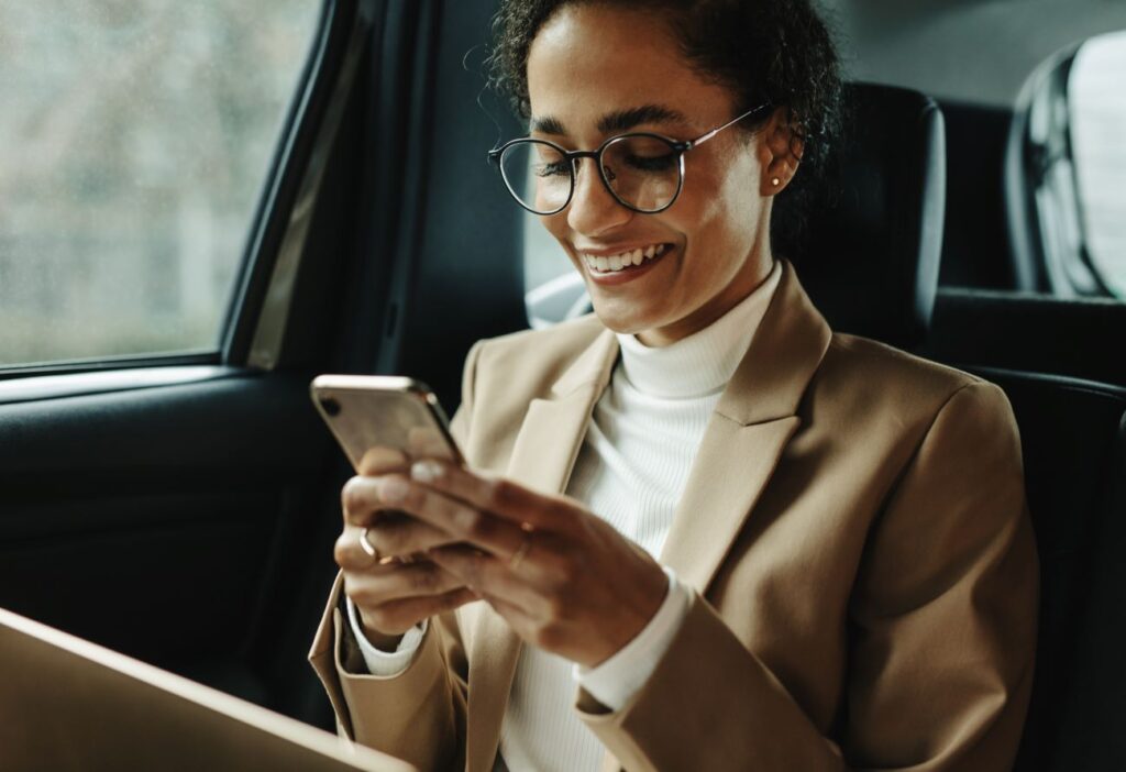 Frau in einem Auto blickt während einer Geschäftsreise lächelnd auf ihr Handy.