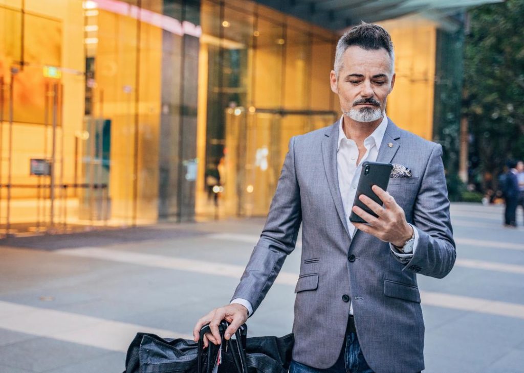 A man in a gray suit looking at his phone on the street while pulling a suitcase.