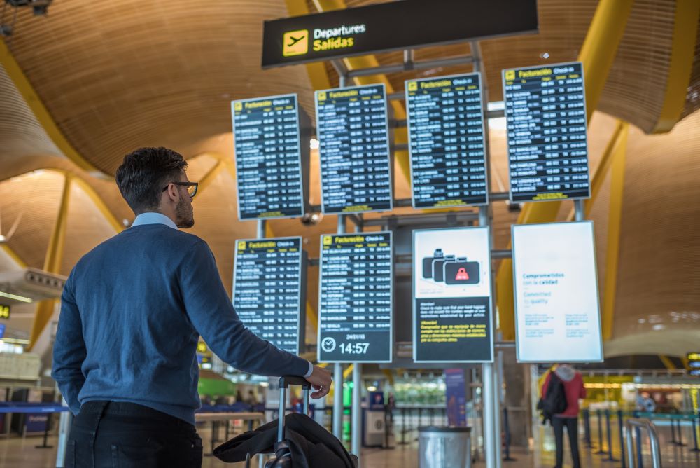 Mann mit Koffer steht vor Anzeigetafel am Flughafen.