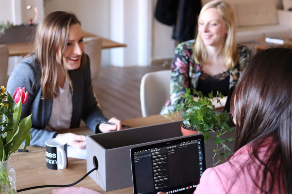 Drei Frauen arbeiten mit Laptops oder Notizen an einem Tisch.