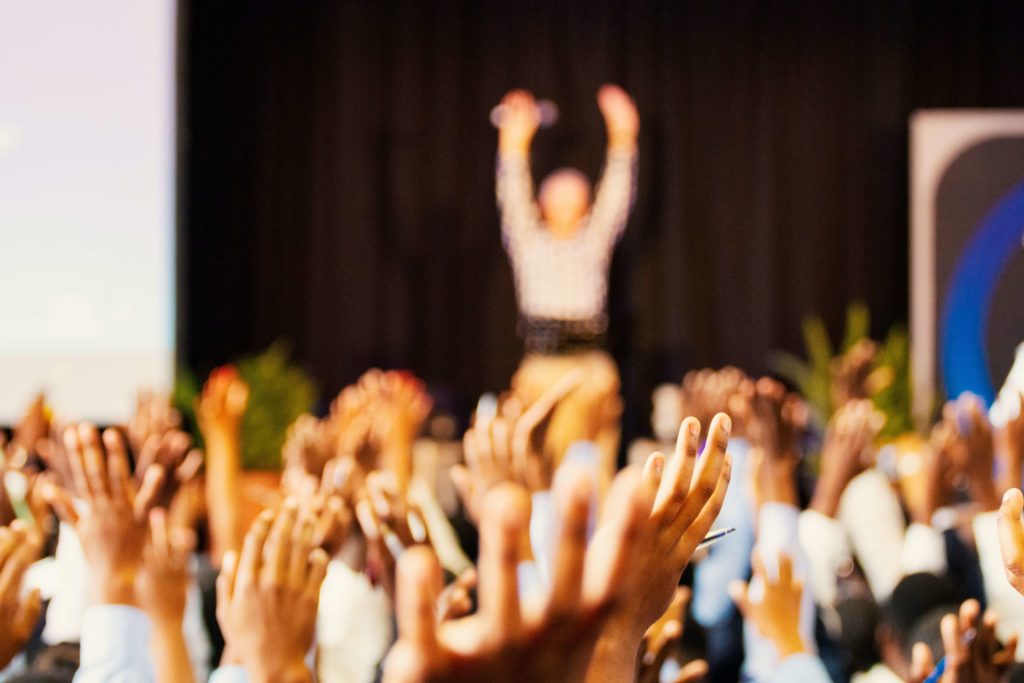 Podiums-Vortrag auf einer Messe. Zuschauer heben Hände. 